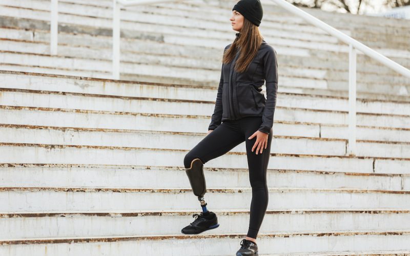 Photo of healthy disabled woman in sportswear with prosthesis leg standing at the stairs and looking aside outdoor